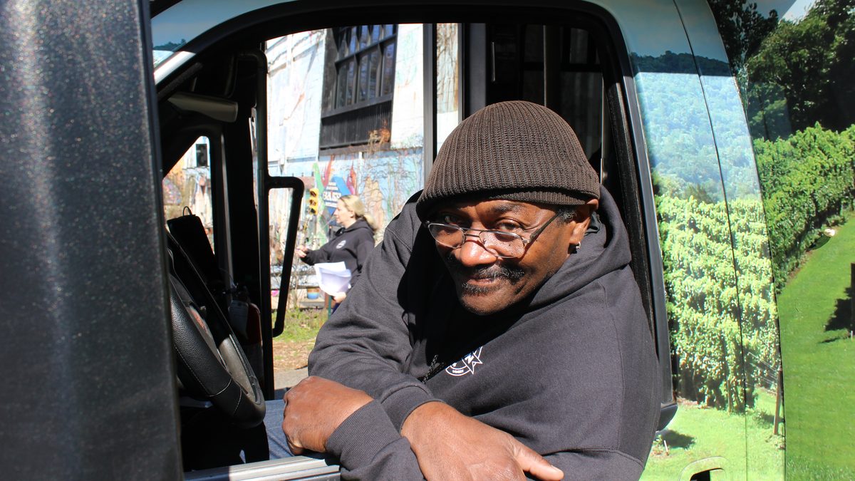 A Hop On driver in a brown beanie and glasses leaning out of the driver's window, smiling, with vineyard art on the bus wrap behind