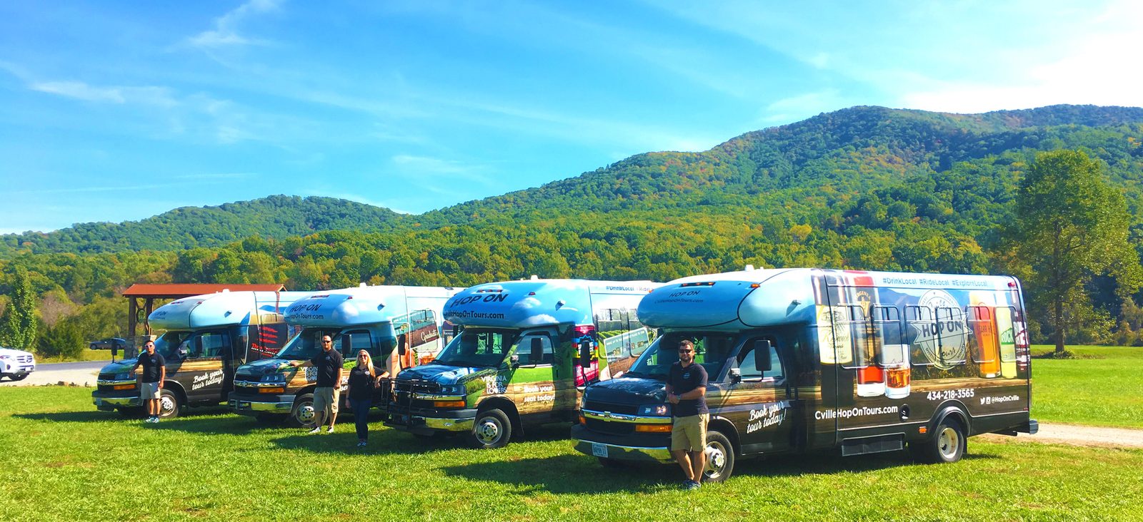 Four Hop On shuttles in their photo wraps parked in a row on a mown field, a driver standing beside each, under a green Blue Ridge ridge and a blue sky