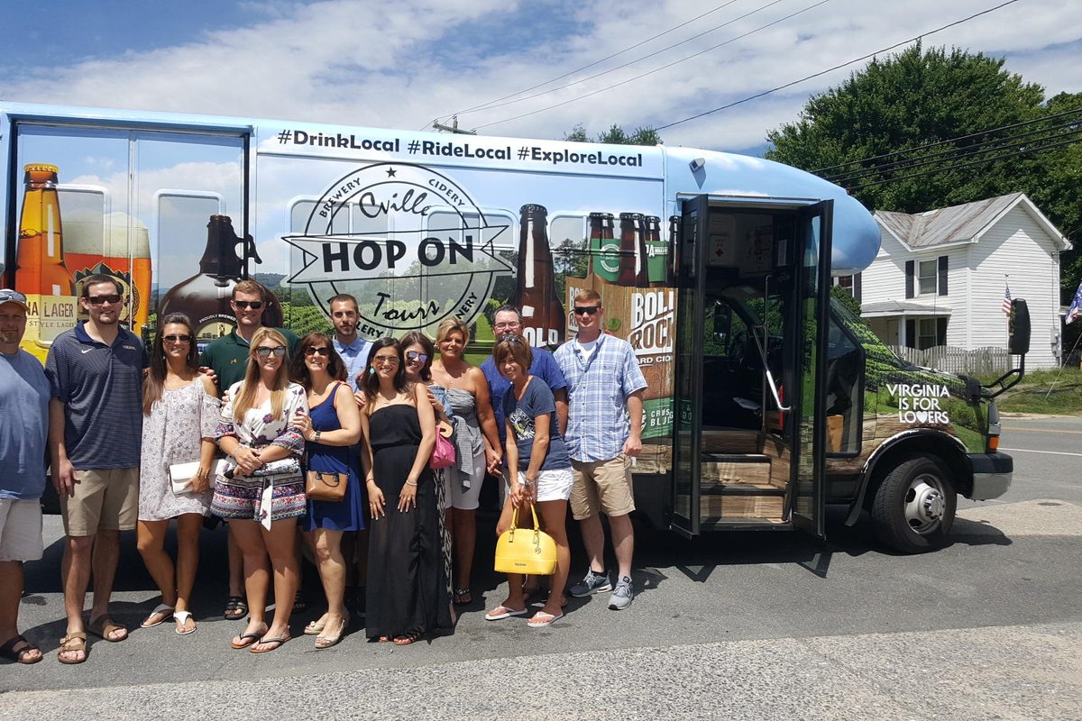 About sixteen riders in summer clothes posed in front of a wrapped Hop On shuttle printed with #DrinkLocal #RideLocal #ExploreLocal and the Hop On roundel