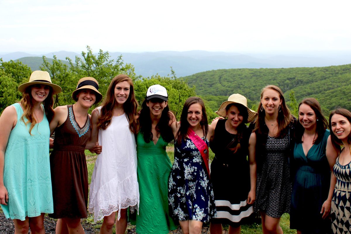 Nine women in sundresses and sun hats arm in arm at a mountain overlook, one wearing a sash