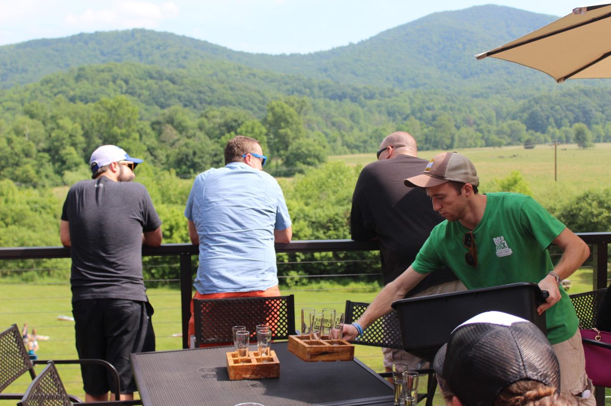 Riders at a deck rail looking over a green valley toward a mountain, tasting flights on the table, a server leaning in