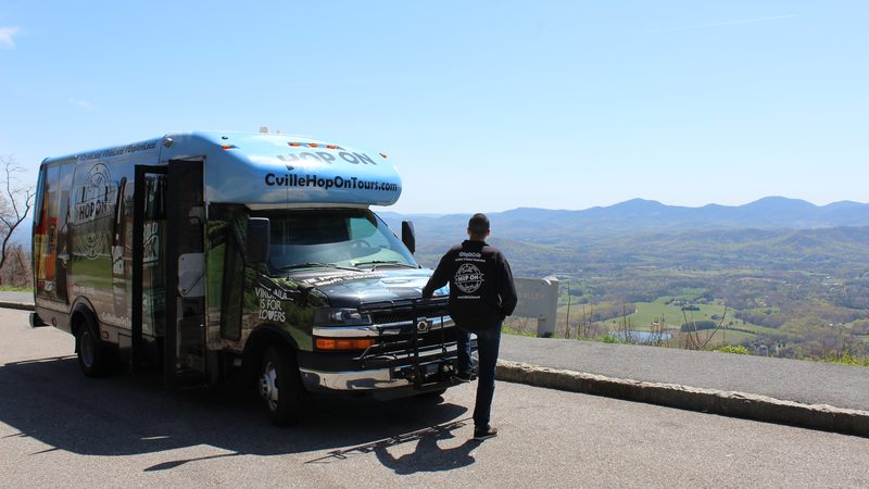A Hop On shuttle parked at a mountain overlook with its door open, and a man in a dark hoodie looking out over the valley